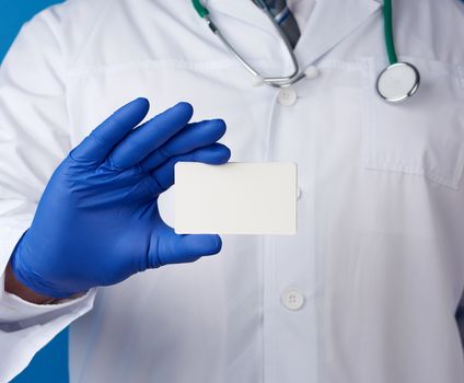 doctor therapist is dressed in a white robe uniform and blue sterile gloves is standing and holding a stack of empty white paper business cards, place for inscription