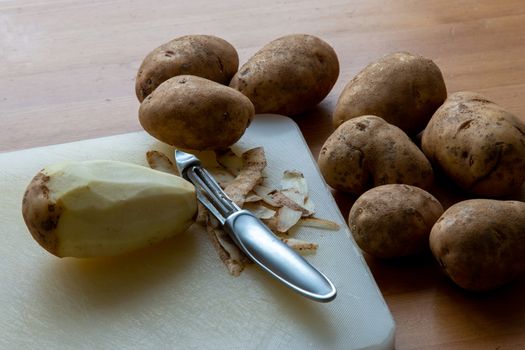A single raw potato lies peeled on a cutting board with a metal peeling tool, among many others still unpeeled.