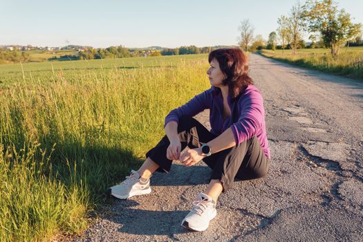 happy european woman resting during running workout on asphalt road on sunset. Attractive and healthy middle age women in countryside.