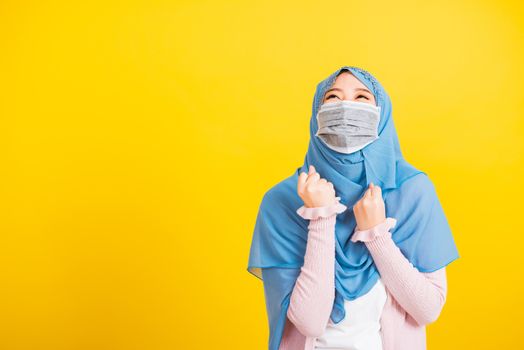 Muslim Arab, Happy beautiful young woman wear veil hijab wear protective face mask quarantines coronavirus raises hands glad excited cheerful after recovering from illness, isolated yellow background