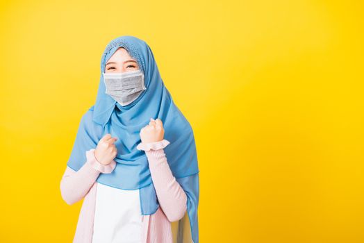 Muslim Arab, Happy beautiful young woman wear veil hijab wear protective face mask quarantines coronavirus raises hands glad excited cheerful after recovering from illness, isolated yellow background