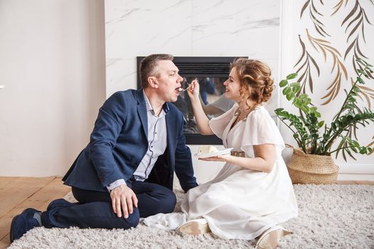 newlyweds. wife feeds her husband with a cake while sitting on the carpet in front of the fireplace indoor