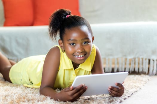 Close up portrait of cute little african girl with ponytail laying on carpet in lounge with tablet.