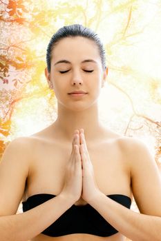Close up conceptual portrait of young woman meditating with tree in background.Girl with eyes closed and hands together in front of chest.