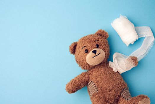 funny vintage brown curly teddy bear with rewound paw with white gauze bandage on blue background, concept of injuries in children or animals, copy space