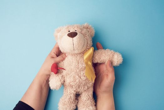 female hands hold a small teddy bear with a yellow ribbon folded in a loop on a blue background. concept of the fight against childhood cancer. problem of suicides