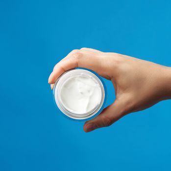female hand holds  a round jar with white cream for face and body on a blue background, concept of moisturizing