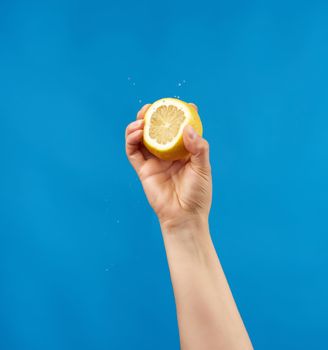 female hand holds half a yellow lemon and squeezes it on a blue background, splashes fly to the sides