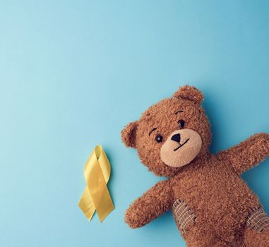 brown teddy bear holds in his paw a yellow ribbon folded in a loop on a blue background. concept of the fight against childhood cancer. problem of suicides and their prevention. copy space
