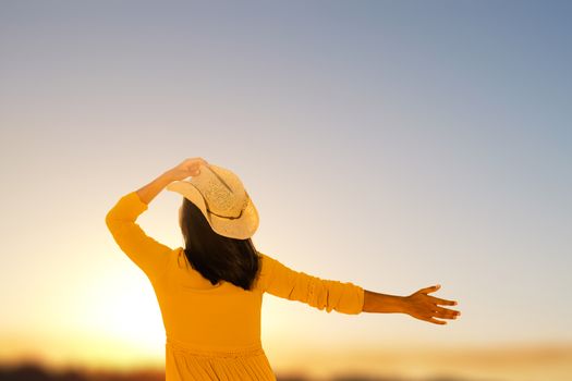 Close up rear view of young woman in yellow dress looking at sunset. Girl holding hat on head with one hand and raising other hand in sky.