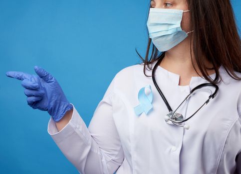 female doctor in a disposable sterile mask and a white coat stands on a blue background and points with the index finger to the side, a blue silk ribbon in the shape of a loop pinned to the chest