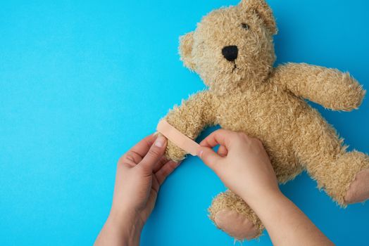 two hands holding a teddy bear and gluing an adhesive plaster on a paw, blue background, top view