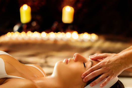 Close up of young woman having healing head massage in spa.Therapist doing manipulative treatment with essential oils on  forehead. Low key scene with candles glowing in background.