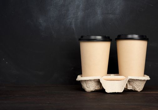 two brown paper disposable cups with a plastic lid stand in the tray on a wooden table, black background, place for an inscription, takeaway containers