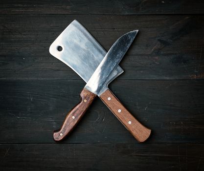 two sharp knives lie on a brown wooden surface, kitchen items are crossed, top view