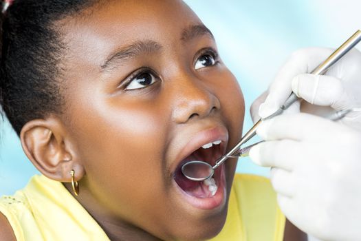 Close up macro portrait of cute little african girl with mouth open having dental health checkup. Dentist examining teeth with hatchet and mouth mirror.