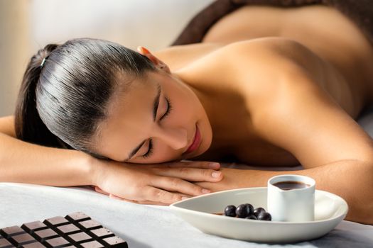 Close up low light portrait of young woman with eyes closed laying on couch in spa. Chocolate slab and hot cacao massage oil next to girl.