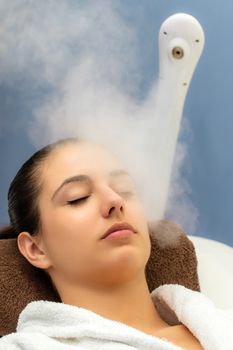 Close up portrait of young woman having thermal steam treatment in spa. Girl with eyes closed in white gown laying on couch.