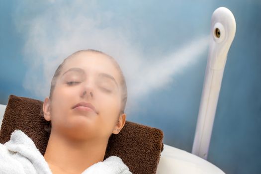 Close up portrait of young woman with eyes closed having thermal steam treatment on face. Girl wearing white gown laying on couch in spa. 