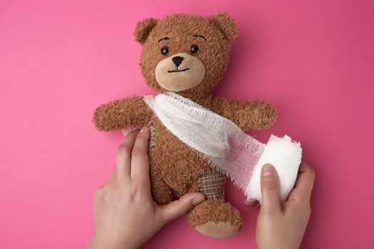 brown teddy bear with bandaged torso with white gauze bandage on a pink background, two female hands are holding a toy