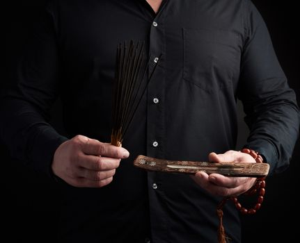 adult man in black clothes holds a stack of incense sticks, dark background