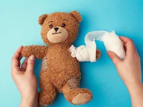 funny vintage brown curly teddy bear with rewound paw with white gauze bandage on blue background, concept of injuries in children or animals, copy space