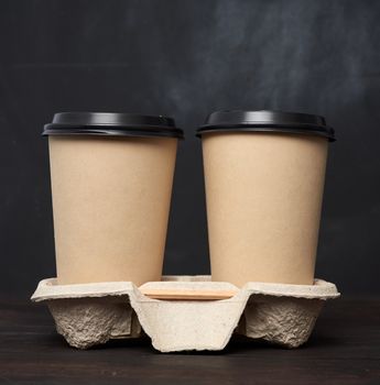 two brown paper disposable cups with a plastic lid stand in the tray on a wooden table, black background, place for an inscription, takeaway containers