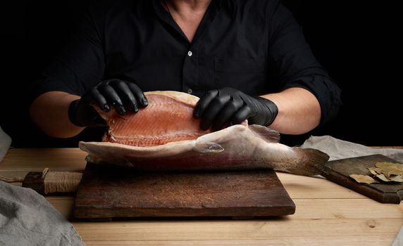 chef in a black shirt and black latex gloves holds a raw carcass of headless salmon fish over a brown wooden cutting board, process of cutting fish