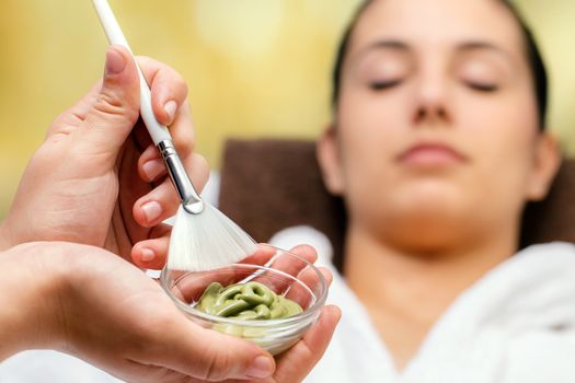 Close up of hands holding bowl and brush with purifying seaweed pealing in spa. Out of focus woman lying on couch in background.