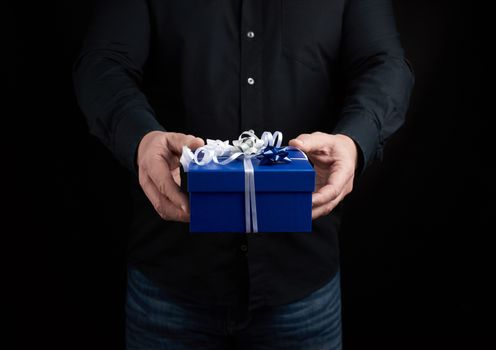 adult man in a black shirt holds a blue square box with a white bow tied on a dark background, concept of congratulations, surprise