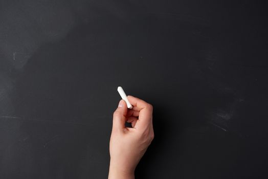 hand holds a piece of white chalk on the background of an empty black chalk board, presentation concept, back to school