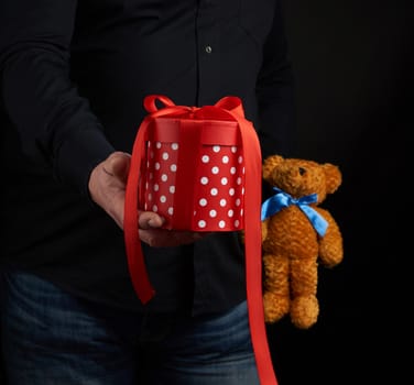 adult man in a black shirt holds a blue square box tied with a red ribbon and brown teddy bear, concept of congratulations on Valentine's Day on February 14, low key 