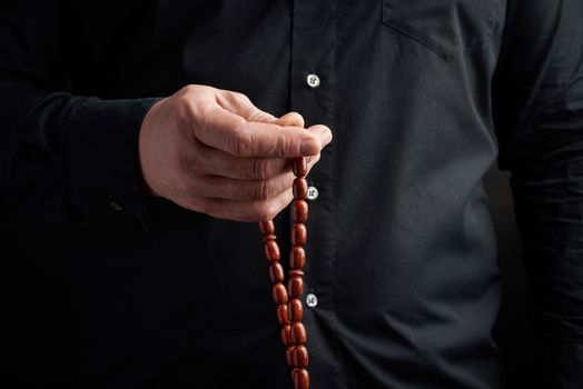 man in a black shirt holds a brown stone rosary in his left hand, low key. used to count prayers or other ritual actions, to preserve attention and concentration, to set the rhythm