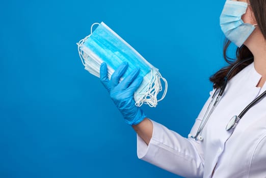 doctor in a white coat and mask holds a stack of protective disposable face masks, physician stands on a blue background, copy space