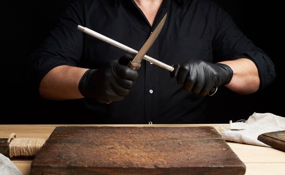 chef in a black shirt and black latex gloves sharpen a kitchen knife on an iron sharpener with a handle above the table, low key