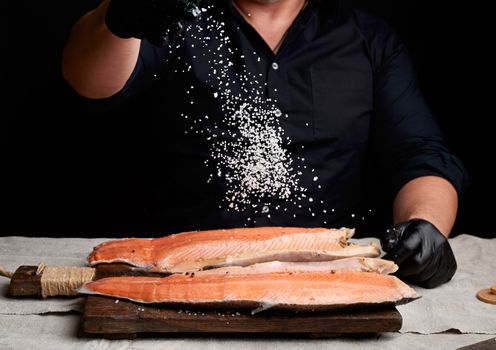 chef in a black shirt and black latex gloves prepares salmon fillet on a wooden cutting board, process of sprinkling with spices and salt, low key