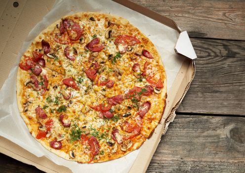 baked round pizza with smoked sausages, mushrooms, tomatoes, cheese and dill in an open cardboard box on a wooden table, food is sliced in portions, top view