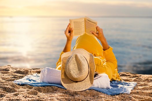 Close up of woman in yellow dress laying on beach reading a book. Rear view of woman wearing hat against idyllic sunset.