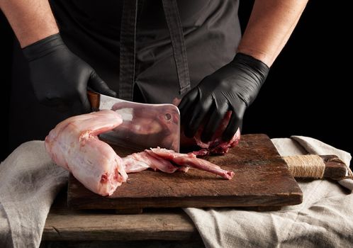 Chef in black latex gloves holds a big knife and cuts into pieces raw rabbit meat on a brown wooden cutting board, cooking on a dark background