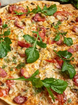 baked round pizza with smoked sausages, mushrooms, tomatoes, cheese and arugula leaves, food is cut in portions