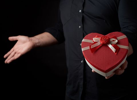 adult man in a black shirt holds a red cardboard box in the form of a heart with a bow on a dark background, concept of holiday, surprise and a gift