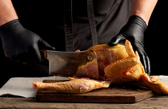 chef in black uniform and latex gloves chopping raw chicken into pieces on a brown wooden board, dark background, cooking process