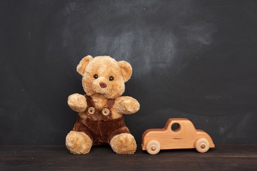 brown teddy bear sits on a brown wooden table and wooden car, behind an empty black chalk board, place for an inscription, back to school