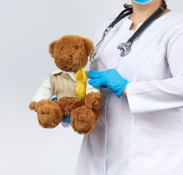 pediatrician in white coat, blue latex gloves holds a brown teddy bear with a yellow ribbon on a sweater, concept of the fight against childhood cancer, problem of suicides