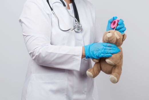 female doctor in a white coat and blue latex gloves holds a brown teddy bear without an eye, doctor wraps his head with adhesive plaster