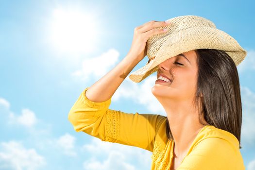 Close up head shot of young carefree woman laughing outdoors. Happy girl in yellow dress holding hat against blue sunny sky.