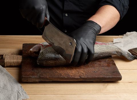 chef in black shirt and black latex gloves slices a whole fresh fish salmon on a vintage brown cutting board,  low key