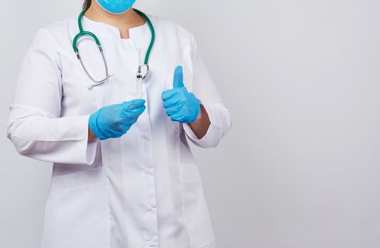 medic woman in white coat and blue latex gloves holds a syringe and shows like gesture with hand, white studio background, concept of timely vaccination against viruses, copy space