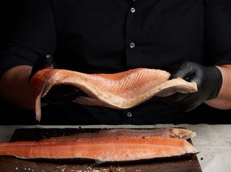 chef in a black shirt and black latex gloves holds a large piece of salmon fish fillet over the table, cooking process, clouse up