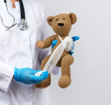 female medic holds brown teddy bear and bandages paw with white gauze bandage, concept of pediatrics and animal treatment